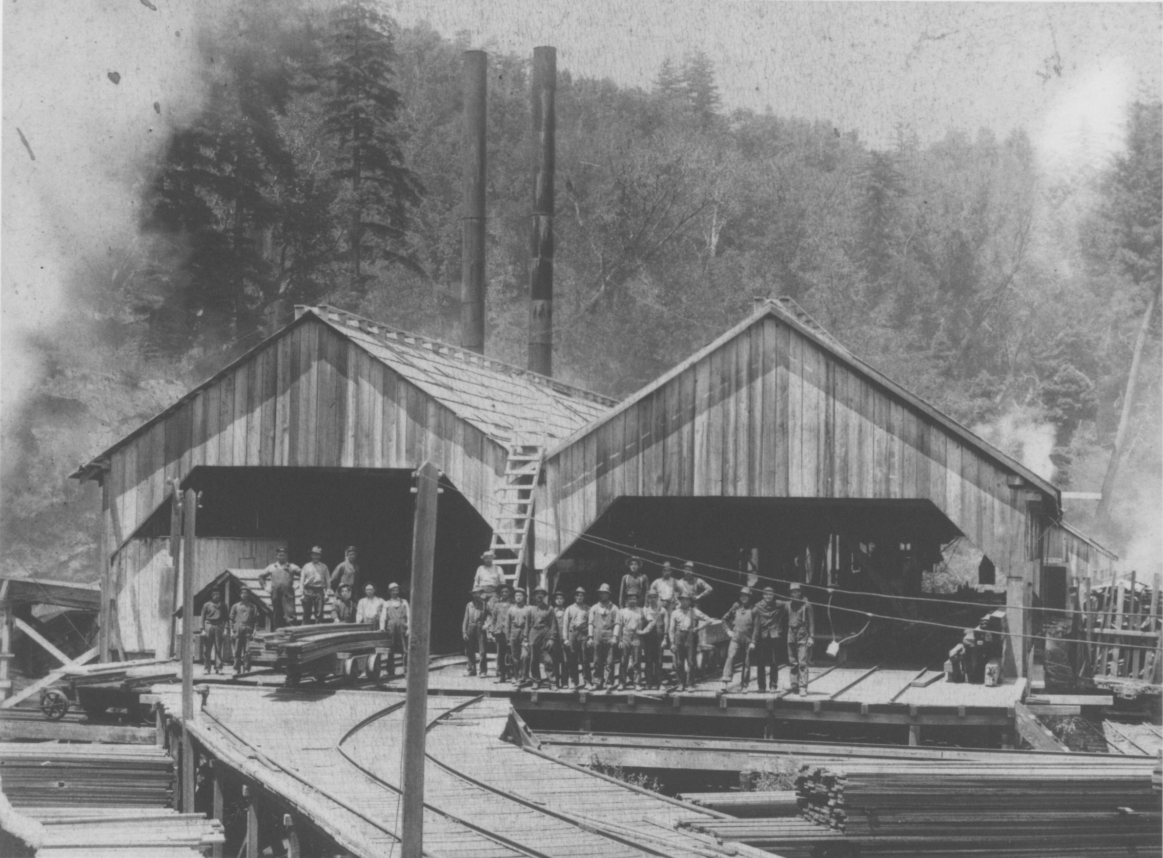 Wooden, peaked roof sheds with open ends and men gathered for the photograph on small scale rail lines at the mill.